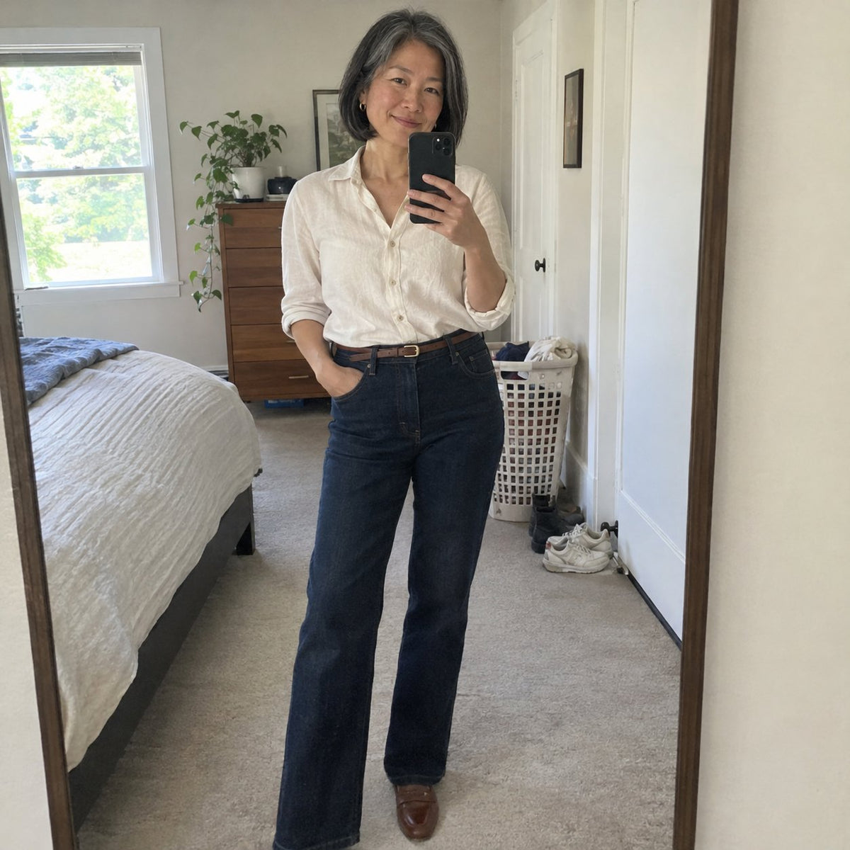 UGC real phone mirror selfie, natural daylight in a tidy bedroom. A 49-year-old woman in tailored dark denim jeans and a tucked-in cream linen blouse, smiling relaxed at her reflection