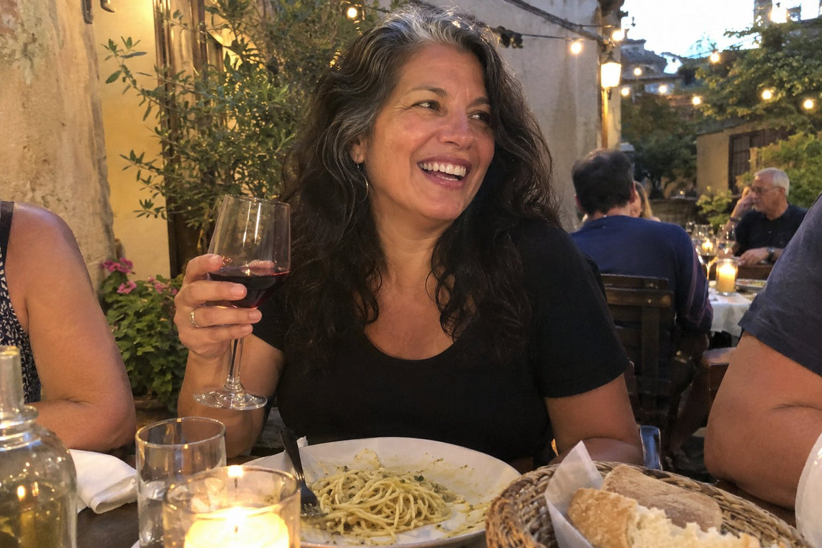 Real candid phone photo at an inviting outdoor Italian trattoria at dusk. A woman in her early fifties laughing warmly across a candlelit table, holding a glass of pinot noir, with a small plate of artisan pasta and oliv