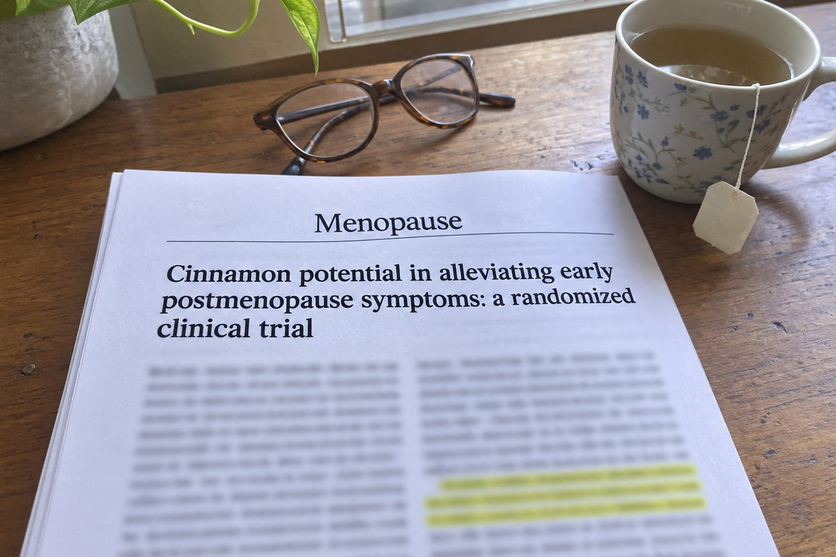 Editorial still-life photo of a printed medical journal paper resting on a wooden desk beside a warm cup of herbal tea. The paper's title "Effects of Cinnamon on Menopausal Symptoms: A Triple-Blind Randomized Controlled 